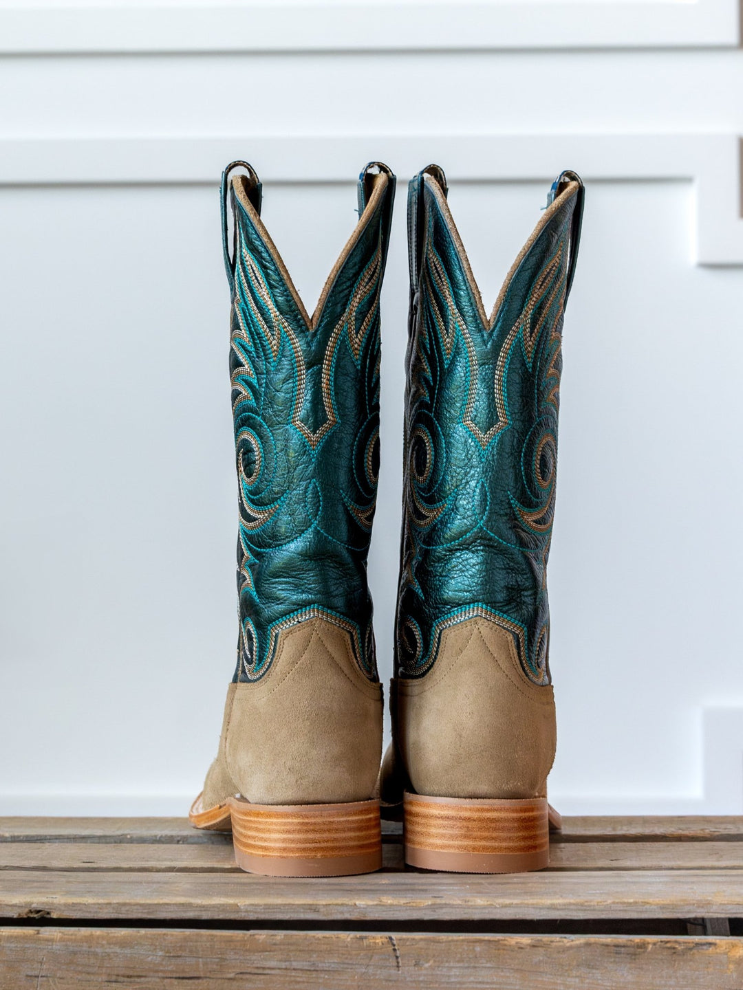 The Salina Metallic Turquoise Boot by Savannah Sevens western life{&}style, featuring tan suede cowhide feet and tall teal blue embroidered shafts, stands side by side on a wooden surface against a white background.