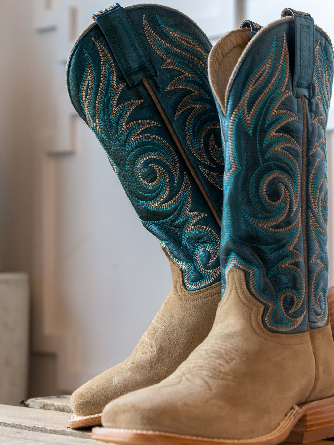 The Hyer Salina Metallic Turquoise Boot by Savannah Sevens western life{&}style features tan leather, square toes, and teal shafts with intricate swirling embroidery, standing upright on a wooden surface against a light background.
