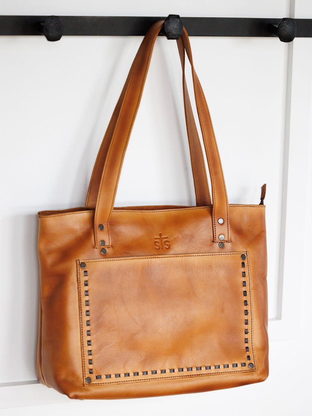 The Saltillo Leather Tote Handbag by Savannah Sevens western life{&}style is a tan leather tote with double shoulder straps, front pocket with metal rivets, subtle embossed logo, and concealed carry holster, shown on a black wall hook against a white background.