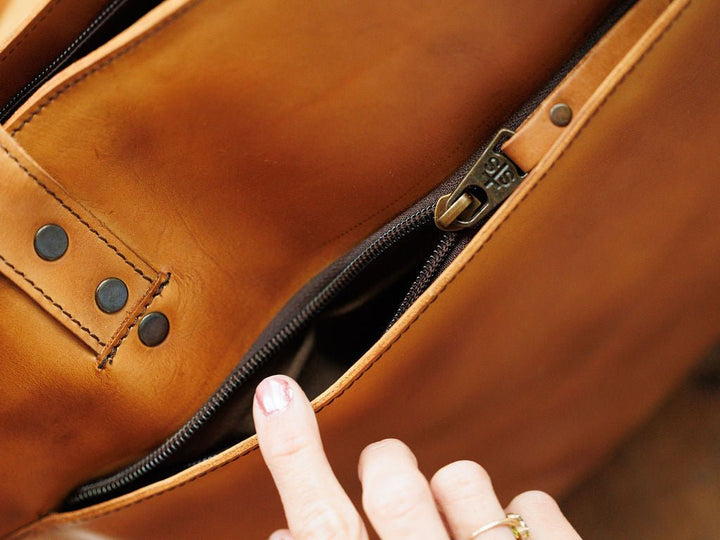 A close-up of the Saltillo Leather Tote Handbag by Savannah Sevens western life{&}style shows brown leather, black rivets, visible stitching, and a hand touching the edge of a partially open zipper for concealed carry space.