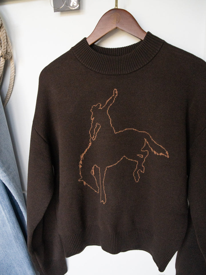The San Antonio Espresso Western Sweater from Savannah Sevens western life{&}style hangs on a wooden hanger, featuring a dark brown knit design with a stitched outline of a person riding a bucking horse. Blue jeans and a rope are partially visible beside it, enhancing the western aesthetic of this graphic sweater.