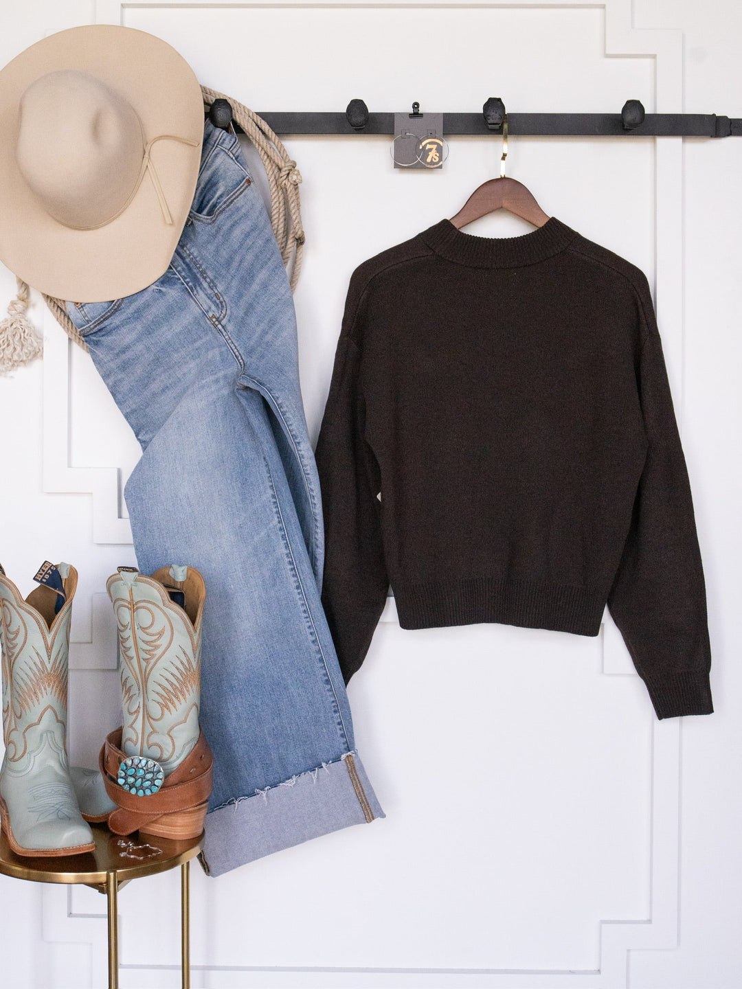 The San Antonio Espresso Western Sweater from Savannah Sevens western life{&}style is displayed alongside a beige cowboy hat, light blue jeans, and a pair of turquoise cowboy boots against a white wall.