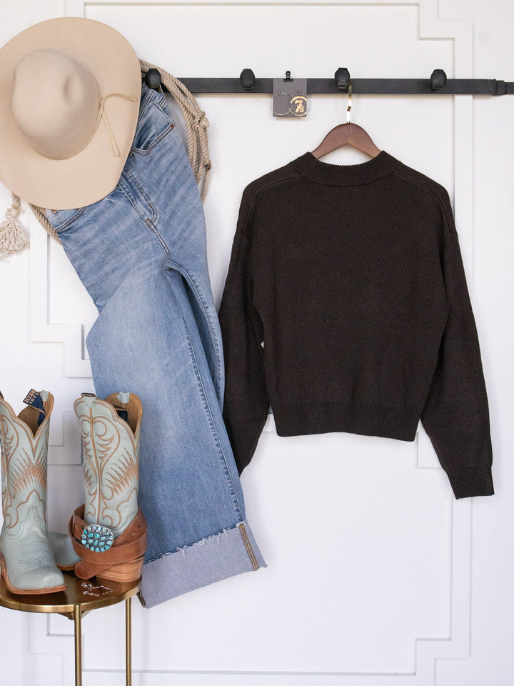 The San Antonio Espresso Western Sweater from Savannah Sevens western life{&}style is displayed alongside a beige cowboy hat, light blue jeans, and a pair of turquoise cowboy boots against a white wall.