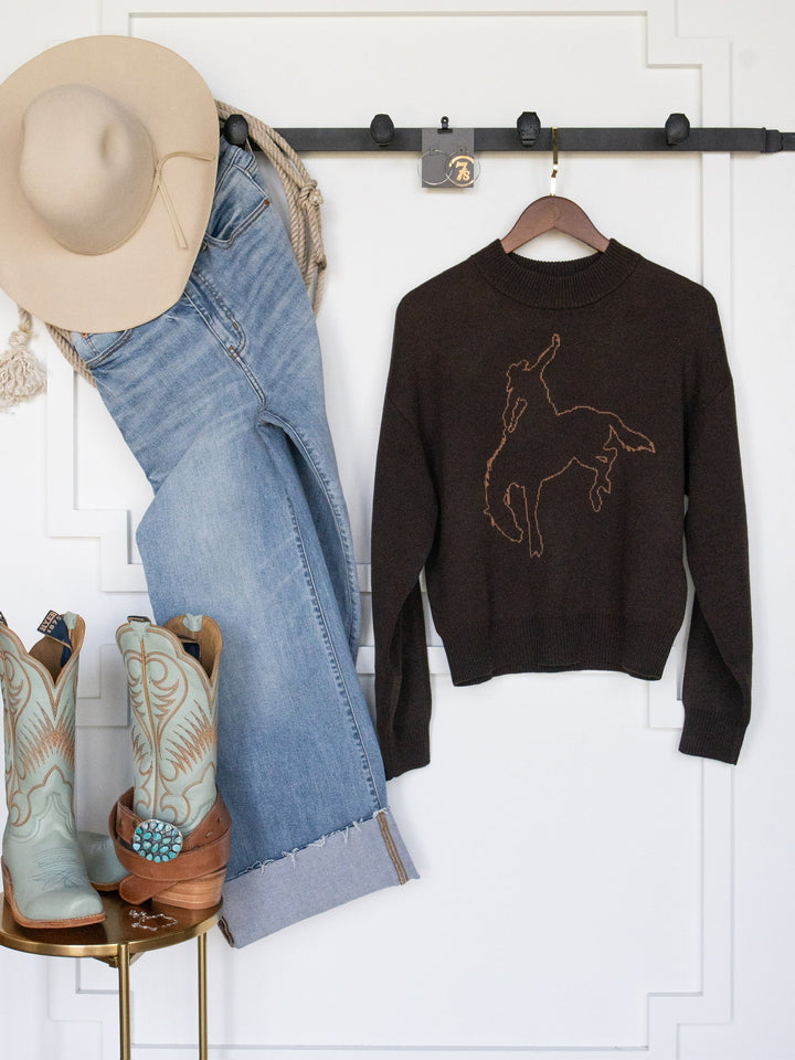 A beige cowboy hat, blue jeans, and turquoise cowboy boots are arranged beside the San Antonio Espresso Western Sweater by Savannah Sevens western life{&}style—a brown long sleeve sweater featuring a rodeo rider graphic—all hanging on a white wall.