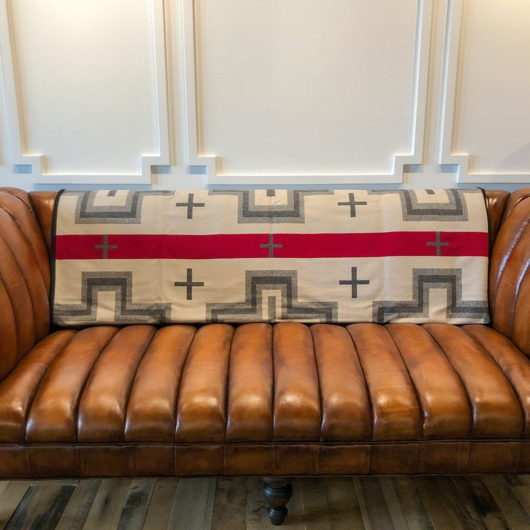 A brown leather sofa with vertical tufting features the San Miguel Blanket by Savannah Sevens western life{&}style, in beige, gray, and red geometric patterns, draped over its backrest against a paneled cream wall.
