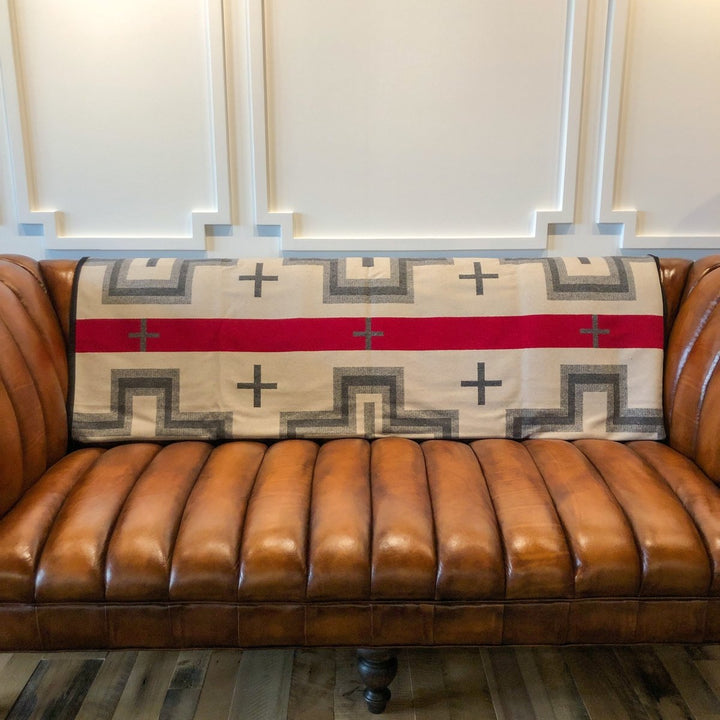 A brown leather sofa with vertical tufting features the San Miguel Blanket by Savannah Sevens western life{&}style, in beige, gray, and red geometric patterns, draped over its backrest against a paneled cream wall.
