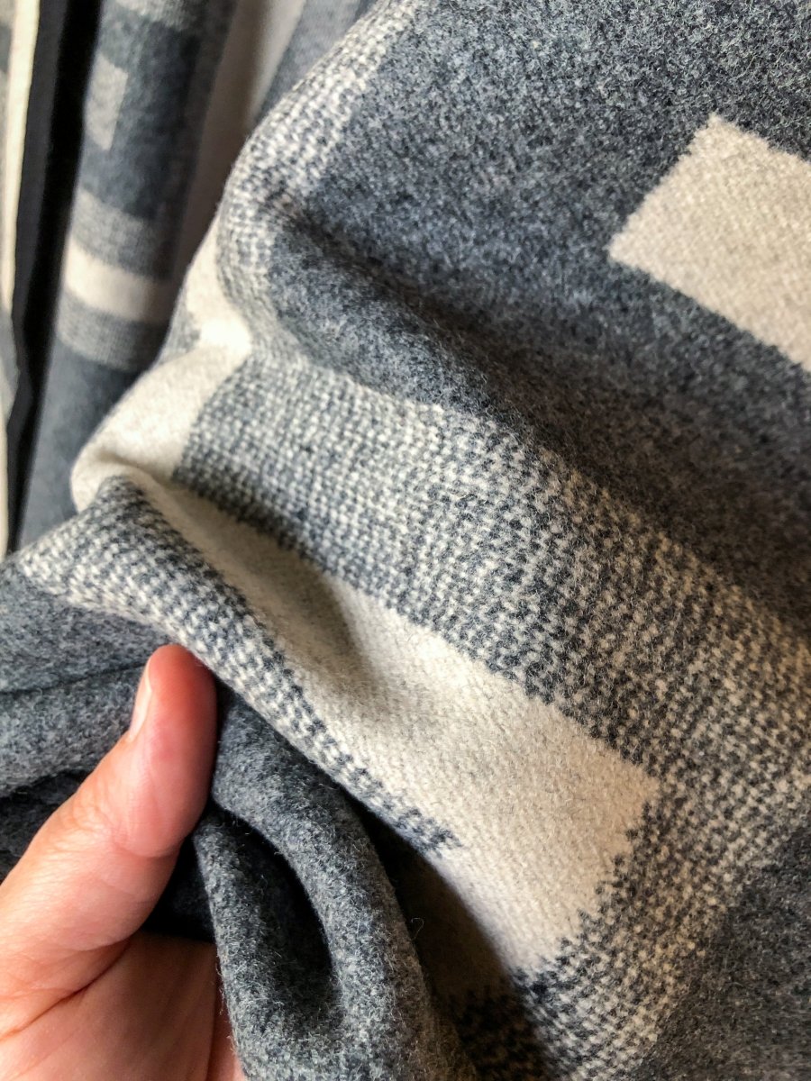 A hand gently holds a soft, gray and white plaid fabric, highlighting the texture and checked pattern of the San Miguel Blanket by Savannah Sevens western life{&}style up close.
