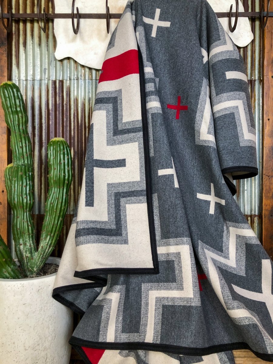 A gray, cream, and red geometric-patterned San Miguel Blanket by Savannah Sevens western life{&}style hangs on a hook beside a tall green cactus in a white pot, set against corrugated metal and wood paneling.