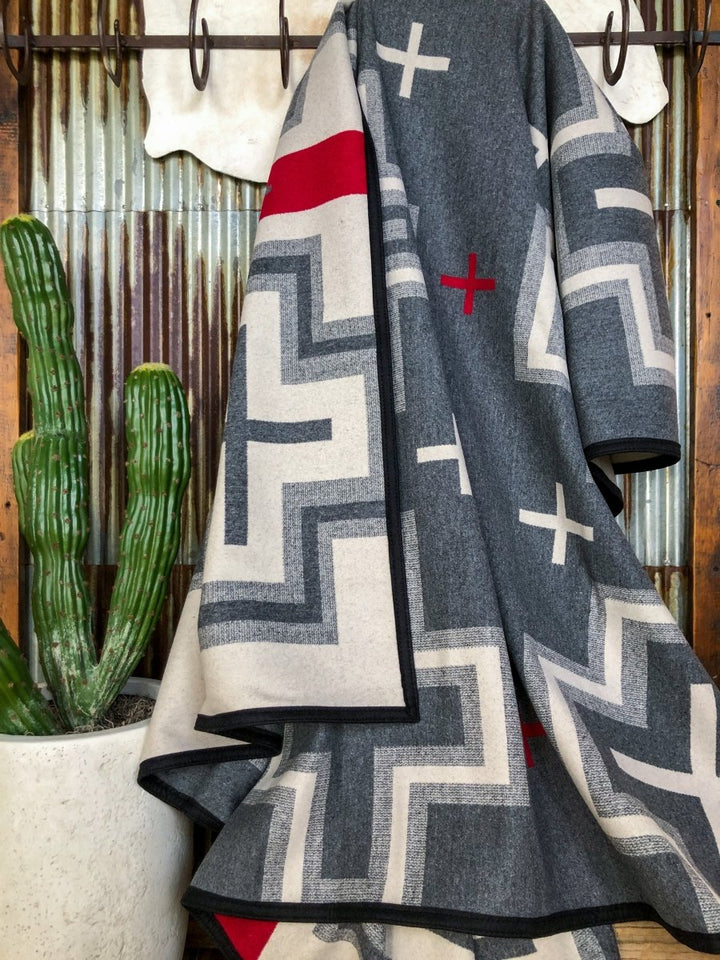 A gray, cream, and red geometric-patterned San Miguel Blanket by Savannah Sevens western life{&}style hangs on a hook beside a tall green cactus in a white pot, set against corrugated metal and wood paneling.