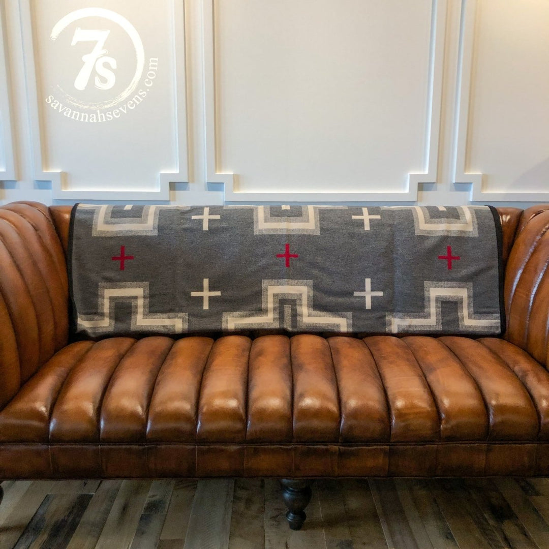 A brown leather tufted sofa with a Savannah Sevens western life{&}style San Miguel Blanket, featuring red and cream plus signs, draped over the backrest. Decorative white paneling adorns the wall behind the sofa.