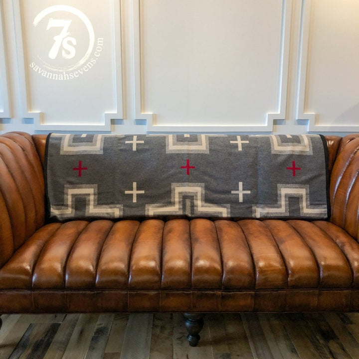 A brown leather tufted sofa with a Savannah Sevens western life{&}style San Miguel Blanket, featuring red and cream plus signs, draped over the backrest. Decorative white paneling adorns the wall behind the sofa.