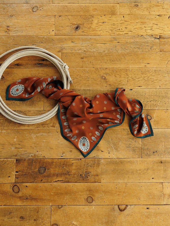 Rust-colored San Miguel Silk Scarf by Savannah Sevens western life&style featuring a green border and repeating geometric print, resting on a wooden floor next to a coiled rope.