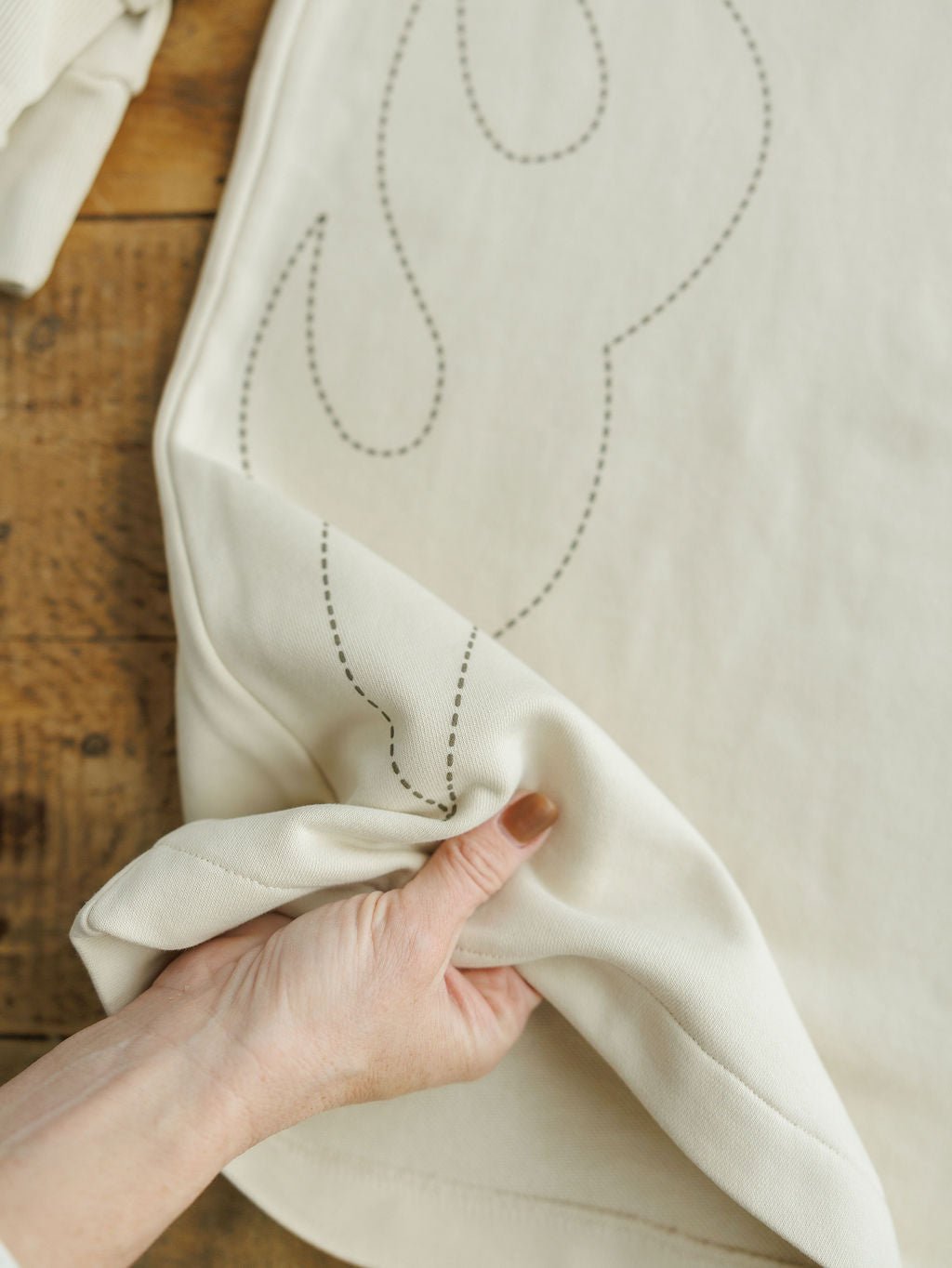 A hand with painted nails lifts the corner of a cream-colored fabric with subtle stitched outlines, displayed on rustic wood—part of the Sanger Natural Boot Stitch Set by Savannah Sevens western life{&}style.
