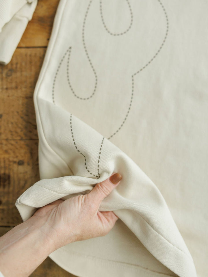 A hand with painted nails lifts the corner of a cream-colored fabric with subtle stitched outlines, displayed on rustic wood—part of the Sanger Natural Boot Stitch Set by Savannah Sevens western life{&}style.