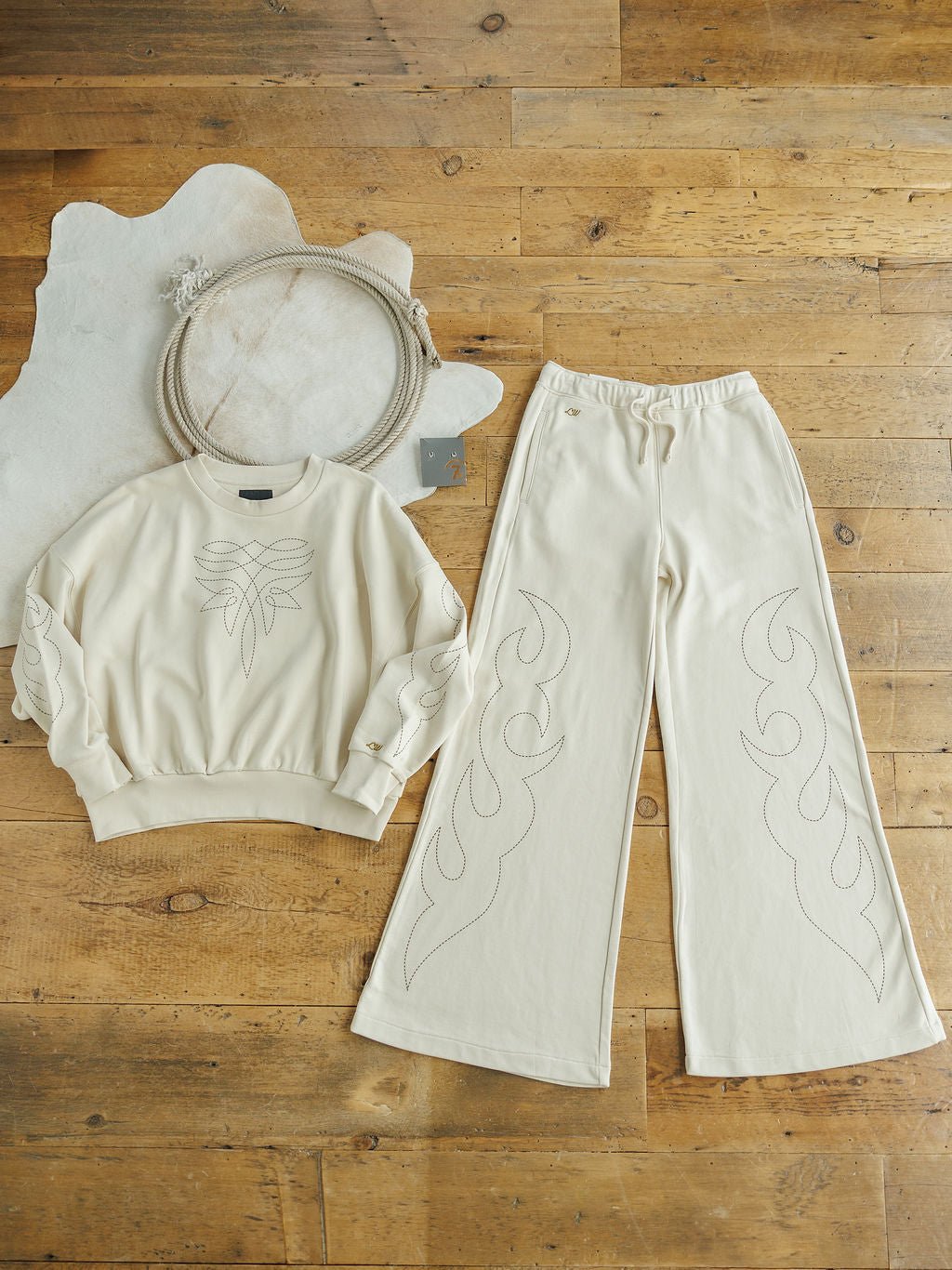 The Sanger Natural Boot Stitch Set by Savannah Sevens western life{&}style features a cream sweatshirt and relaxed, high-waisted pants with subtle boot stitch embroidery, styled beside rope and a hide rug from the Lainey Wilson Collection.