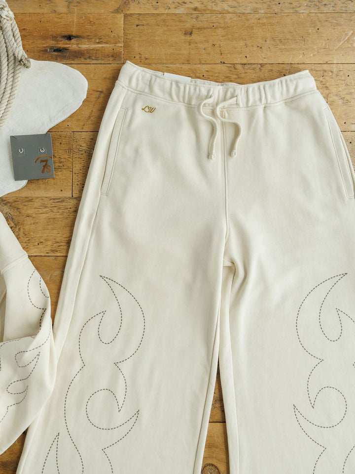 The Sanger Natural Boot Stitch Set by Savannah Sevens western life{&}style features cream high-waisted pants with stitched, wavy flame-like designs, shown on wood. A matching sweatshirt sleeve and a tagged rope complete the Lainey Wilson Collection look.