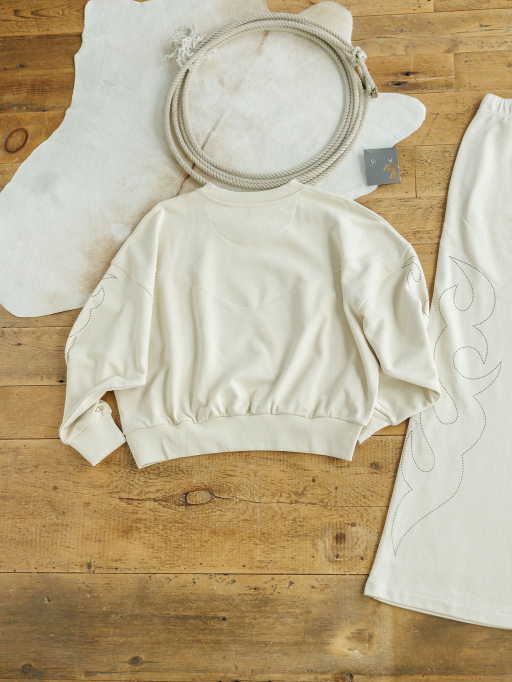 The Sanger Natural Boot Stitch Set from Savannah Sevens western life{&}style features a cream sweatshirt and matching skirt with stitched designs, laid out on a wooden floor for a cozy, relaxed fit perfect for any casual day.