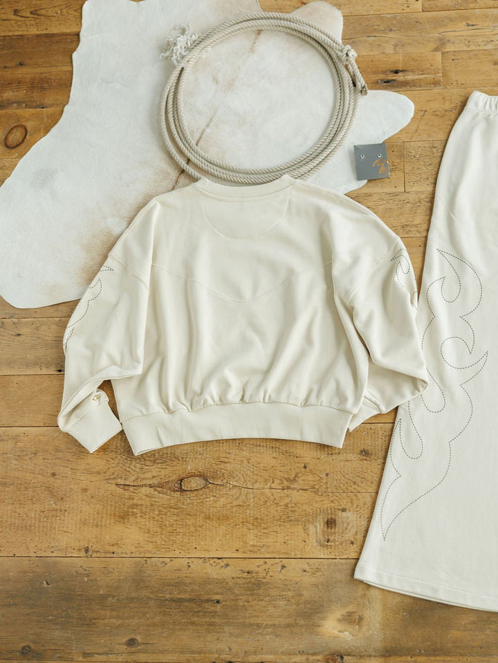 The Sanger Natural Boot Stitch Set from Savannah Sevens western life{&}style features a cream sweatshirt and matching skirt with stitched designs, laid out on a wooden floor for a cozy, relaxed fit perfect for any casual day.
