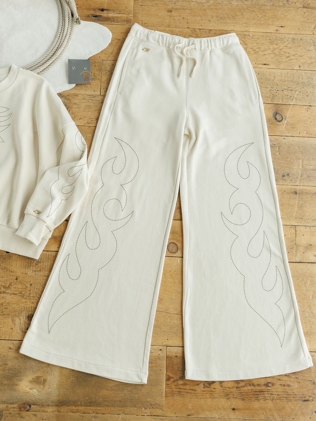 The Sanger Natural Boot Stitch Set from Savannah Sevens western life{&}style features cream-colored high waisted pants with a relaxed fit and subtle flame stitching, shown on a wooden floor beside the matching sweatshirt.