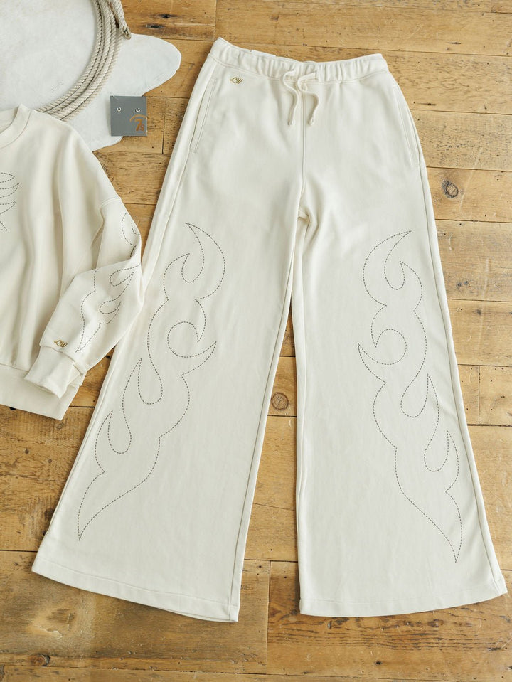 The Sanger Natural Boot Stitch Set from Savannah Sevens western life{&}style features cream-colored high waisted pants with a relaxed fit and subtle flame stitching, shown on a wooden floor beside the matching sweatshirt.