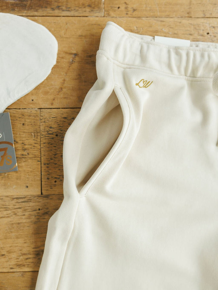 Close-up of ivory high-waisted pants with relaxed fit and side pocket, featuring gold embroidered LW initials near the waistband on a wooden surface. Part of the Sanger Natural Boot Stitch Set by Savannah Sevens western life{&}style.
