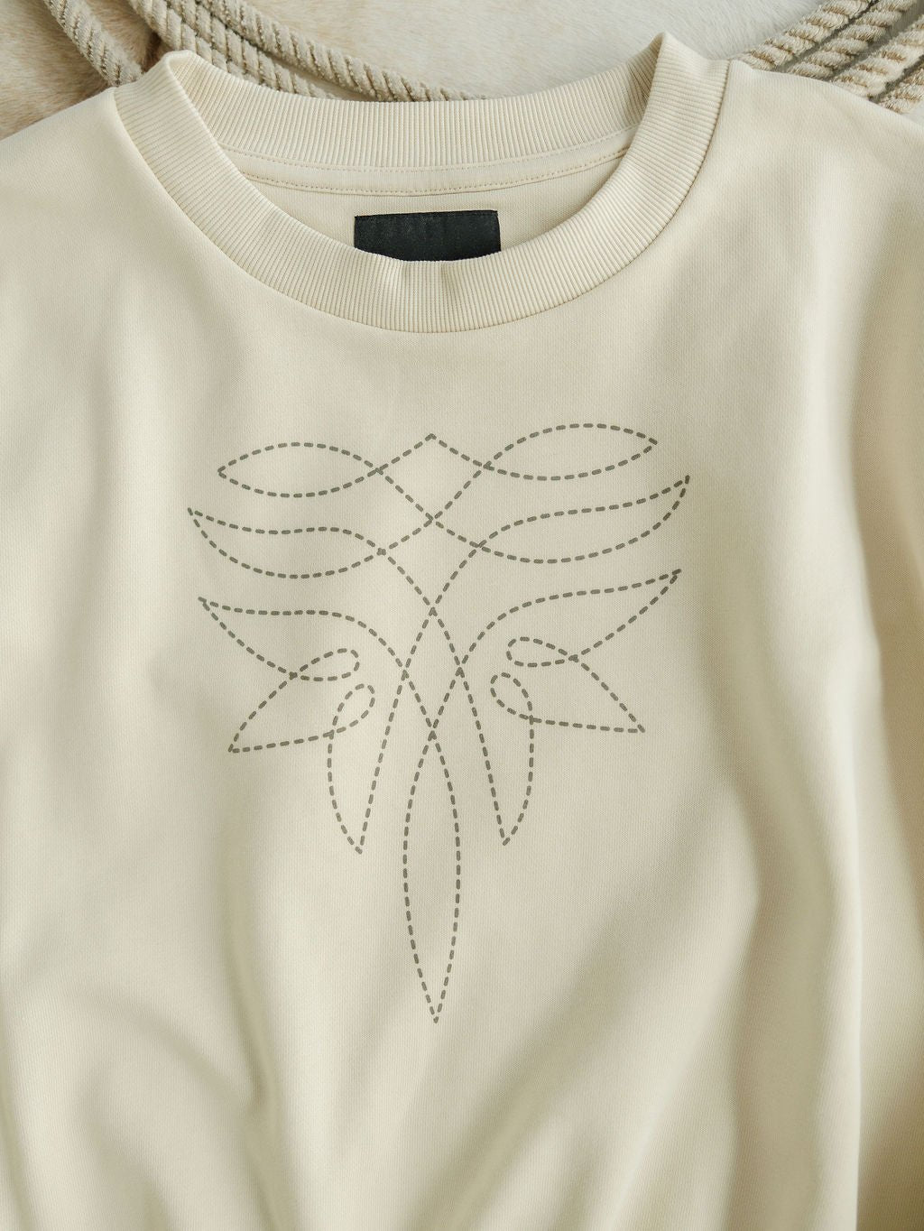 The Sanger Natural Boot Stitch Set by Savannah Sevens western life{&}style is a cream-colored sweatshirt featuring a dotted, abstract geometric front design. It offers a relaxed fit, ribbed neckline, and has a black label inside the collar.