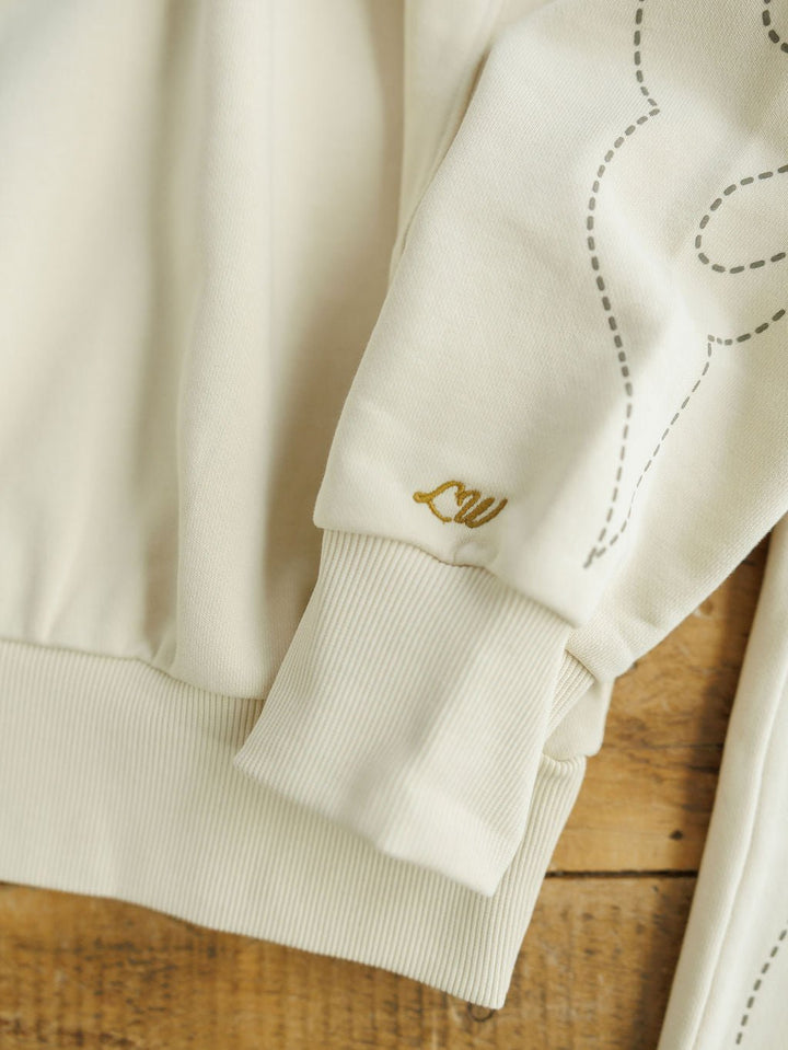 Close-up of a cream-colored Sanger Natural Boot Stitch Set sweatshirt sleeve with ribbed cuffs, gold embroidered script, and partial dashed outline, by Savannah Sevens western life{&}style, laid on a wooden surface.