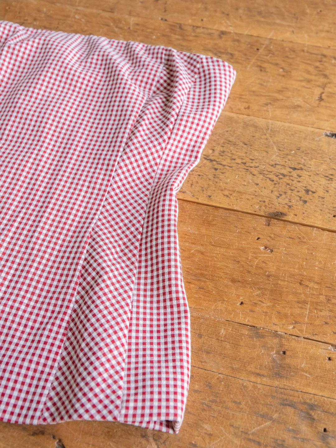 The Sanibel by Savannah Sevens western life{&}style features a red and white small checkered cotton blend fabric, reminiscent of gingham, shown partially folded on a wooden surface.