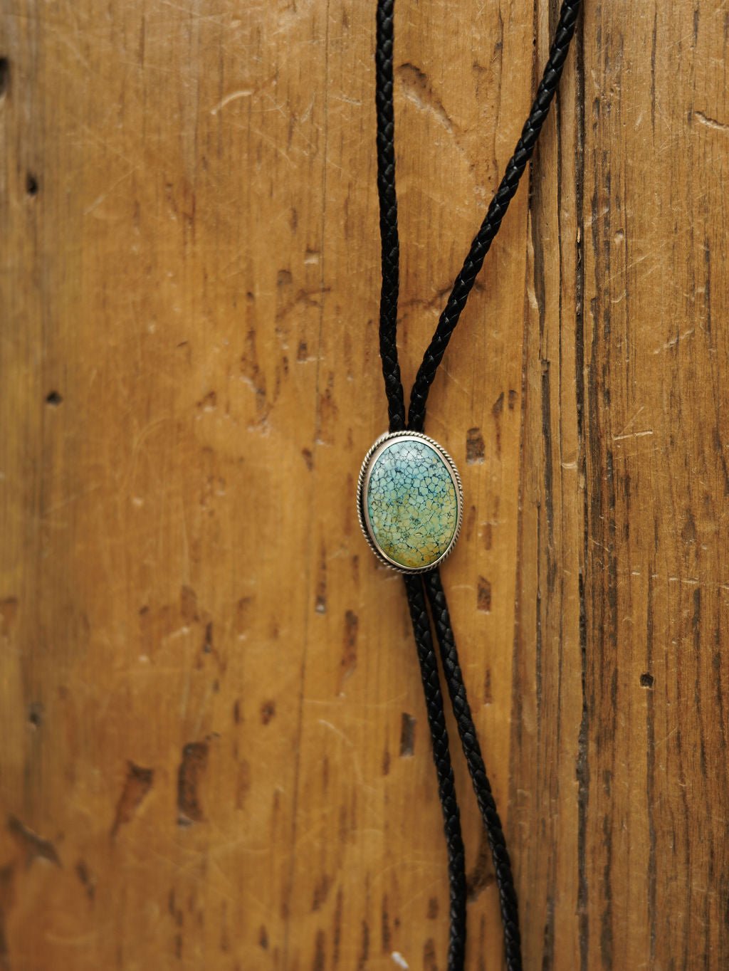 A close-up of the Santa Fe Bolo by Savannah Sevens western life{&}style, featuring a braided black leather cord and an oval Cloud Mountain Turquoise stone clasp set in sterling silver, shown on a rustic wooden background.