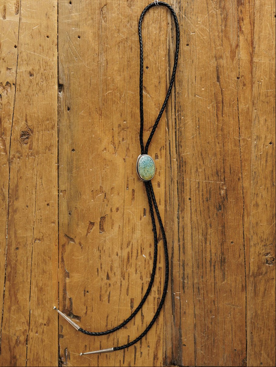 The Santa Fe Bolo by Savannah Sevens western life{&}style features a black braided leather cord with sterling silver tips and an oval Cloud Mountain Turquoise clasp, displayed on rustic wood.