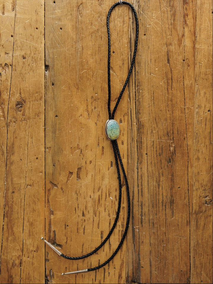 The Santa Fe Bolo by Savannah Sevens western life{&}style features a black braided leather cord with sterling silver tips and an oval Cloud Mountain Turquoise clasp, displayed on rustic wood.