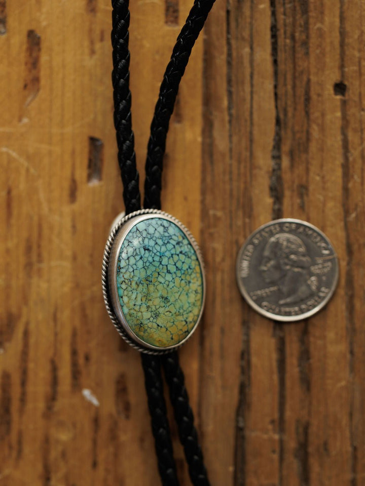 The Santa Fe Bolo by Savannah Sevens western life{&}style features braided black leather and an oval Cloud Mountain Turquoise clasp set in sterling silver, shown next to a U.S. quarter on a wooden surface. The turquoise displays a crackled blue-green pattern.