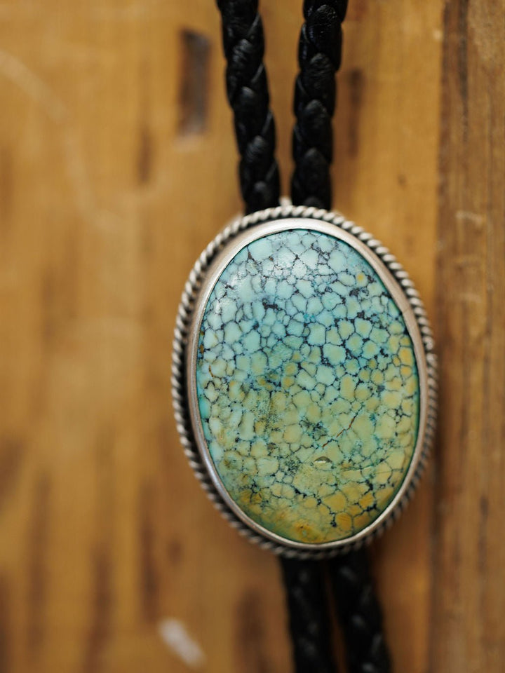 A close-up of the Santa Fe Bolo by Savannah Sevens western life{&}style, featuring an oval Cloud Mountain Turquoise with a crackled pattern set in sterling silver on braided black leather cords, shown against a wooden background.