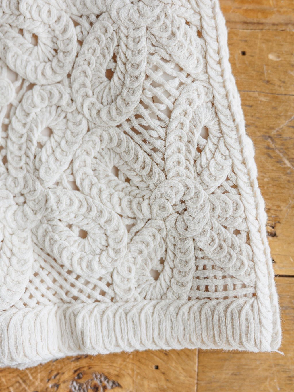 Close-up of The Saratoga by Savannah Sevens western life{&}style, a white macrame textile featuring intricate braided floral and geometric patterns, displayed on a rustic wooden surface beside cream shorts.
