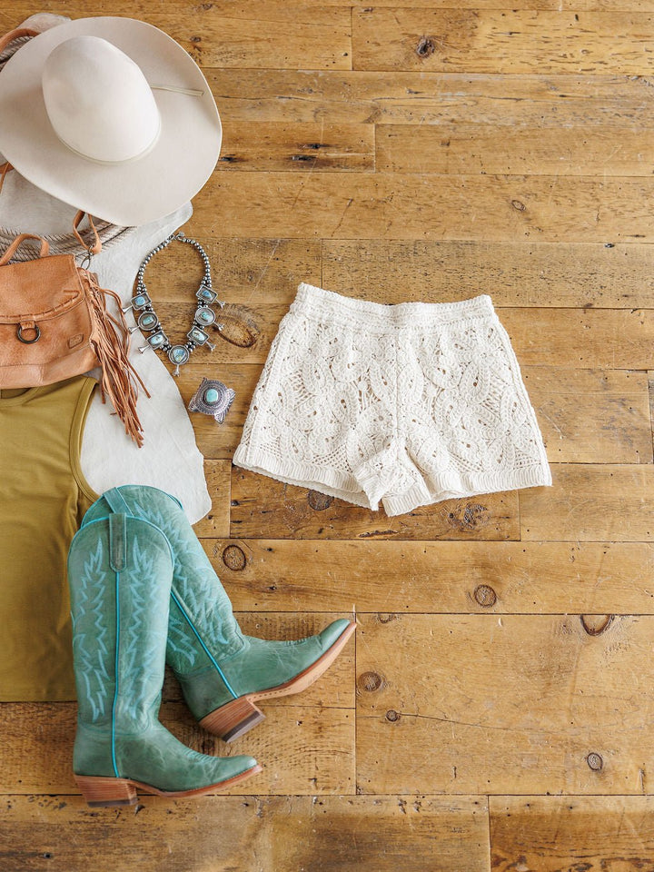 Laid out on a wooden floor, The Saratoga outfit by Savannah Sevens western life{&}style features a pale hat, fringe purse, turquoise necklace and ring, olive shirt, high-waisted cream crochet shorts, and turquoise cowboy boots.