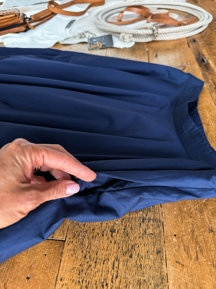 A hand pinches the fabric of the Sarcoxie Navy Skirt by Savannah Sevens western life{&}style, displayed on a wooden table. In the background, white and tan accessories—including ropes and straps—offer perfect companions for styling with this high waisted or full length cotton skirt.