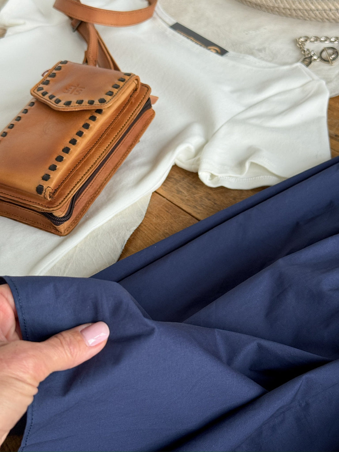 A hand displays the Sarcoxie Navy Skirt by Savannah Sevens western life{&}style near a white shirt, a brown leather purse, and a beige woven bag with a silver chain, all arranged on a wooden surface. This ensemble offers an effortlessly chic look when styled with the high-waisted cotton skirt.