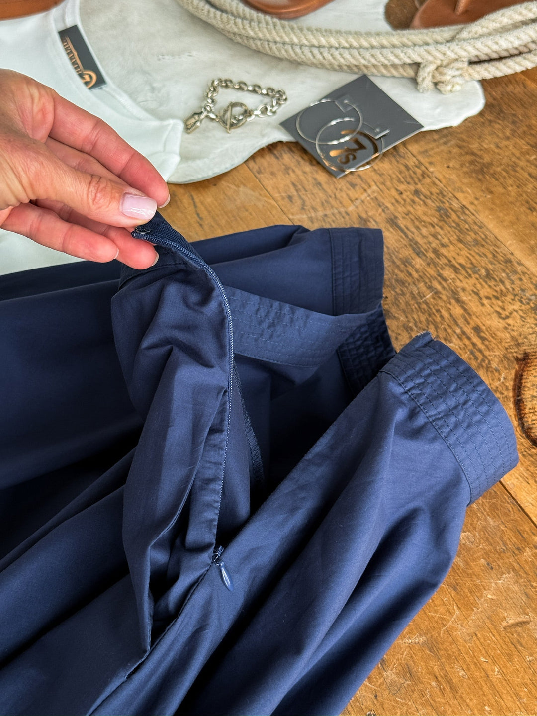 A hand unzips the side of the Sarcoxie Navy Skirt by Savannah Sevens western life{&}style on a wooden table, with a white shirt, necklace, and tan sandals nearby.