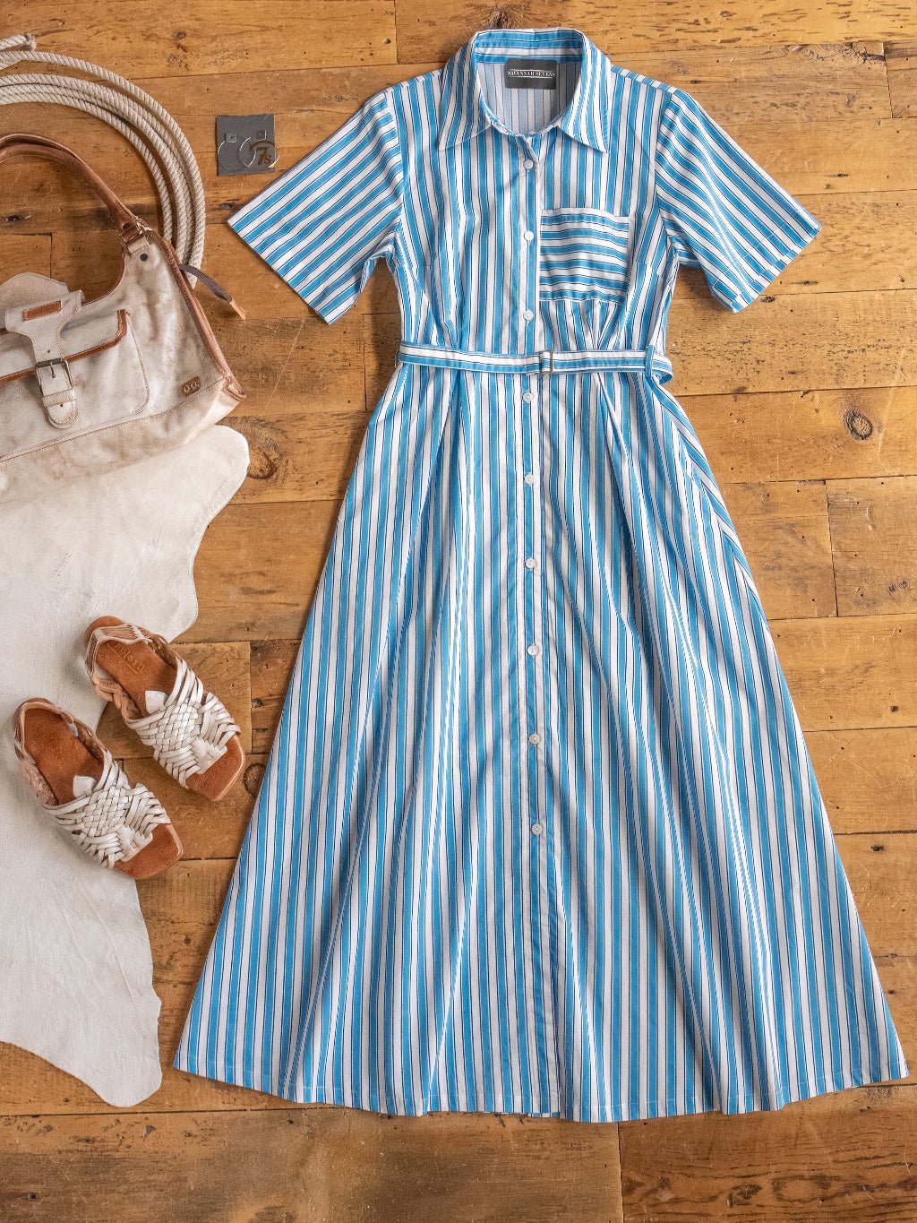 The Sartell Blue Striped Button Up Dress by Savannah Sevens western life{&}style is styled on a wooden floor with a white handbag and braided sandals atop a white cowhide rug.