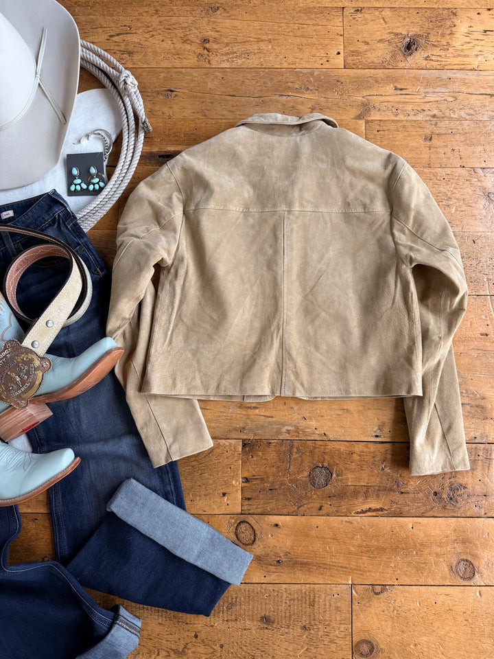 The Sausalito Tan Suede Jacket from Savannah Sevens western life{&}style is laid flat on a wooden floor, surrounded by a white cowboy hat, turquoise earrings, leather boots, jeans, and a belt with a decorative buckle. This women’s suede jacket brings timeless western charm to any outfit.