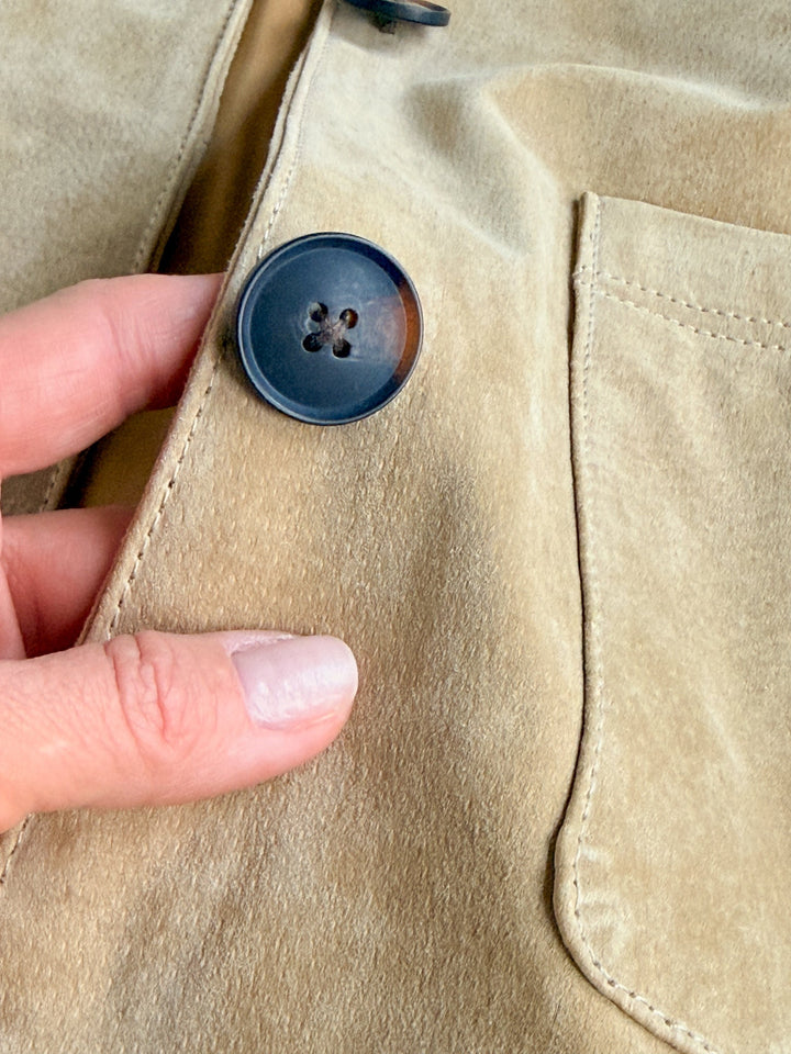 A hand with light pink nails holds the edge of the Sausalito Tan Suede Jacket by Savannah Sevens western life{&}style near a large, dark button and a stitched pocket.
