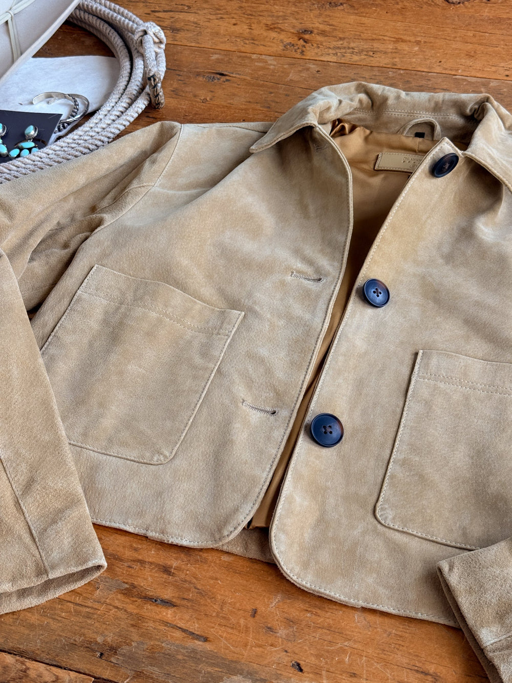 The Sausalito Tan Suede Jacket by Savannah Sevens western life{&}style is laid flat on a wooden surface, showcasing its large front pockets and dark buttons. A section of braided rope and a white purse appear in the top left corner.