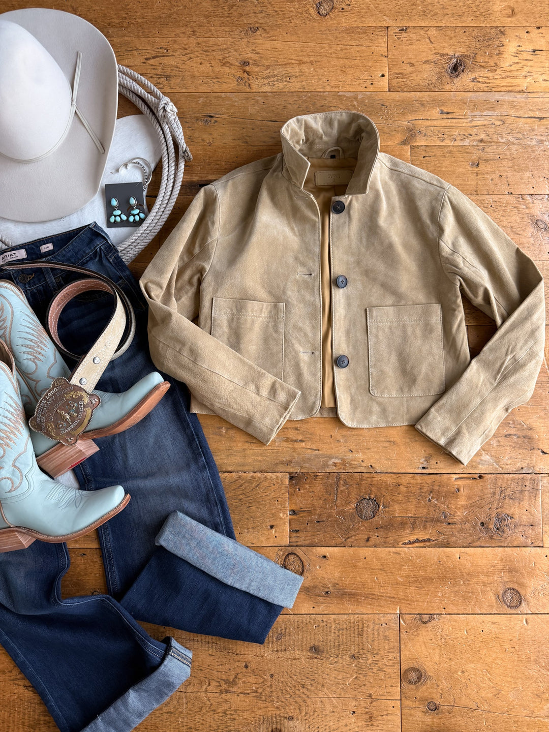 Flat lay of the Sausalito Tan Suede Jacket by Savannah Sevens western life{&}style, paired with blue jeans, turquoise cowboy boots, a white cowboy hat, turquoise earrings, and a woven bag arranged on a wooden floor.
