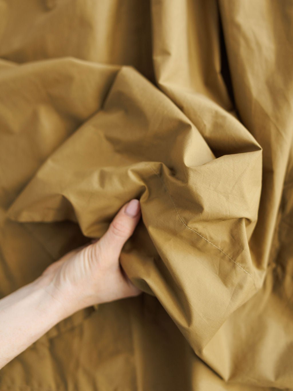 A light-skinned hand gently scrunches soft fabric, creating folds that evoke the comfort and style of the Saville Bronze Dress by Savannah Sevens western life{&}style—a sleeveless midi dress with side pockets.
