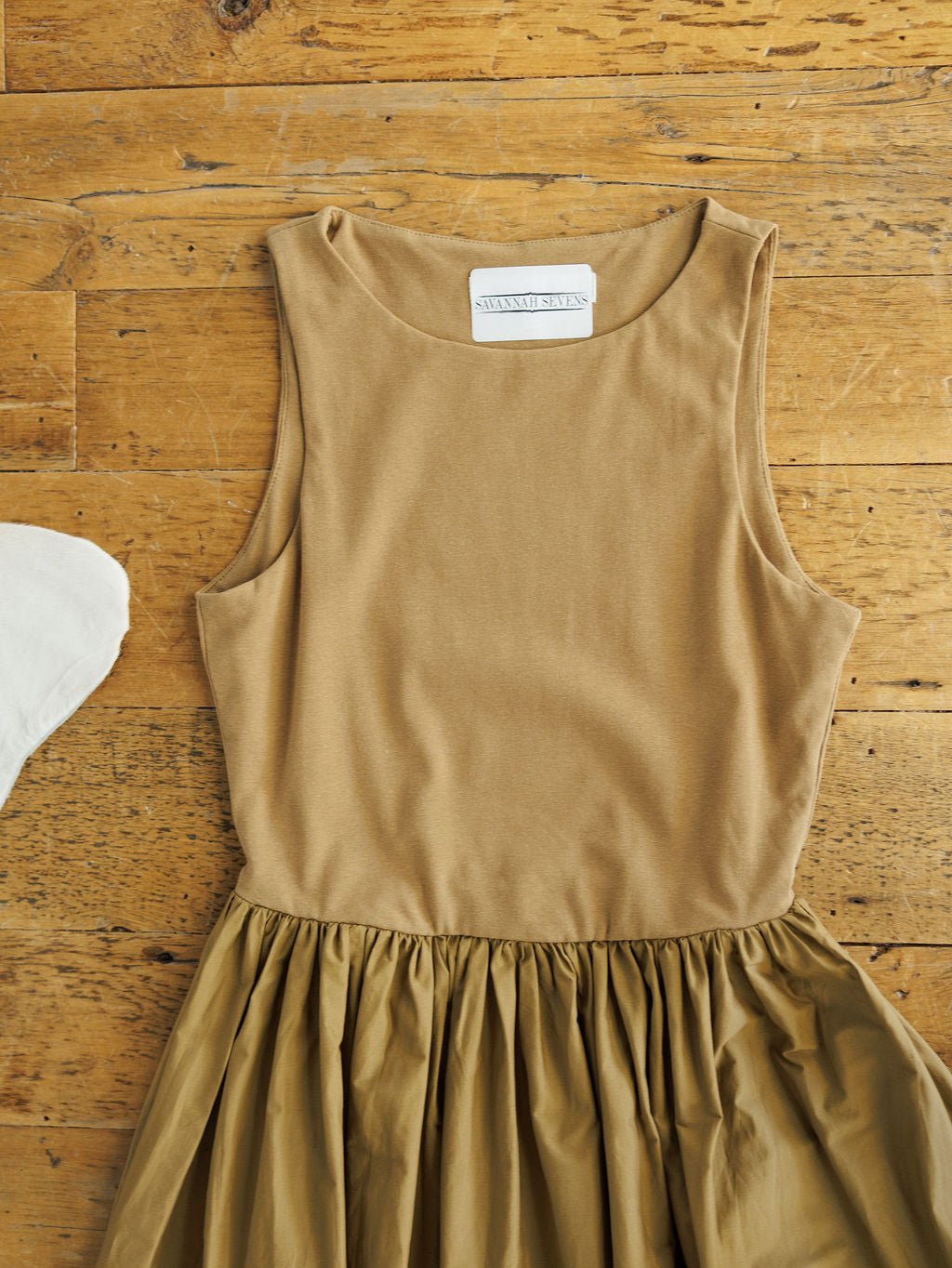 The Saville Bronze Dress by Savannah Sevens western life{&}style, a sleeveless tan cotton midi with fitted top, gathered skirt, and side pockets, is laid flat on rustic wood showing its neckline label.