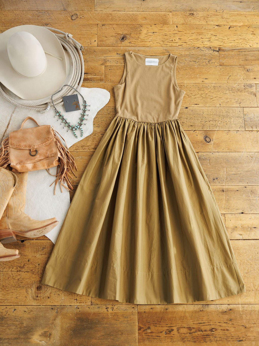 The Saville Bronze Dress by Savannah Sevens western life{&}style, a sleeveless tan cotton midi with a fitted bodice, side pockets, and a full skirt, is styled on wood with a white hat, beaded necklaces, fringed bag, cowboy boots, and cowhide rug.