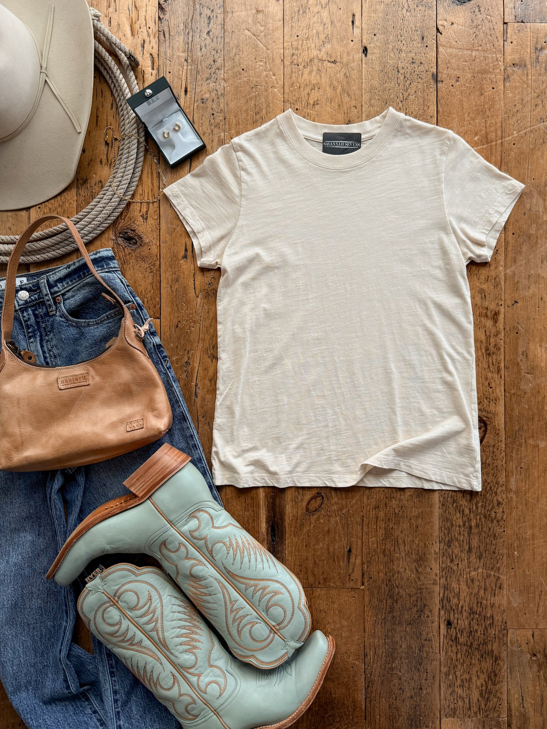 Flat lay of a relaxed fit cream slub Savannah Sevens western life&style Searcy Natural Basic Tee, blue jeans, a tan leather handbag, and light blue western boots, with a cowboy hat and small earrings nearby.