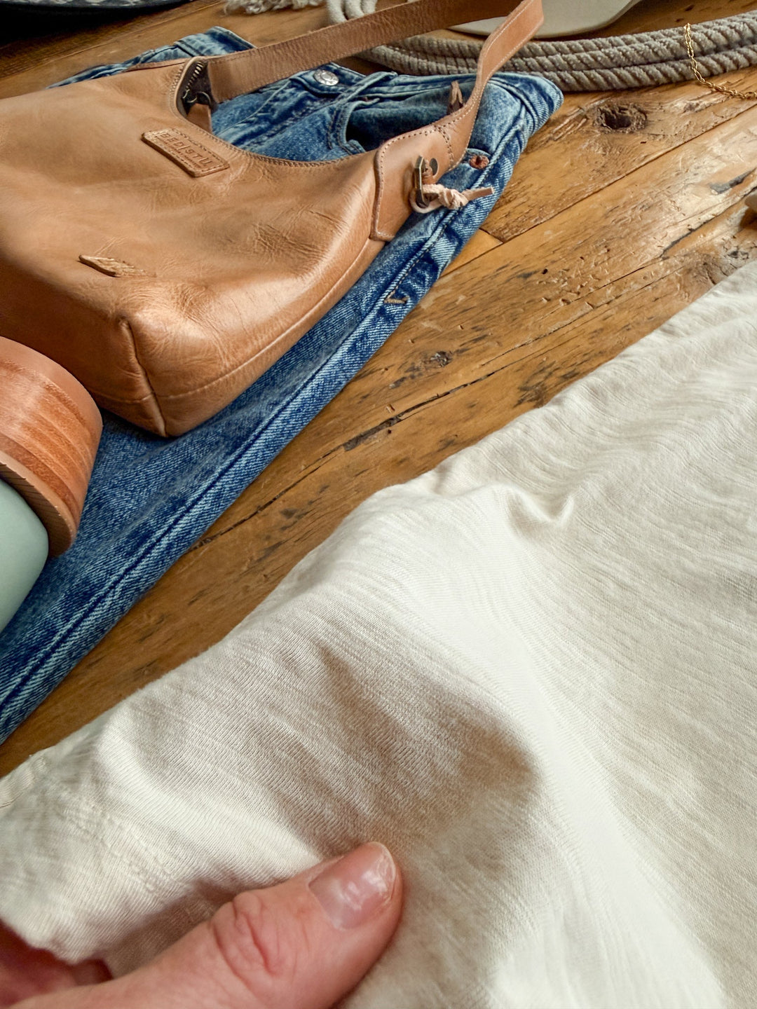 A hand holds a relaxed fit Searcy Natural Basic Tee next to a pair of blue jeans and a tan leather handbag on a wooden surface.
