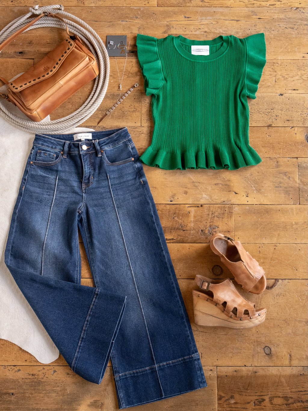 A flat lay displays the Sebring Emerald Flutter Sleeve Top by Savannah Sevens western life{&}style, paired with wide-leg blue jeans, tan platform sandals, a brown shoulder bag, coiled rope, and silver jewelry on a wooden floor.