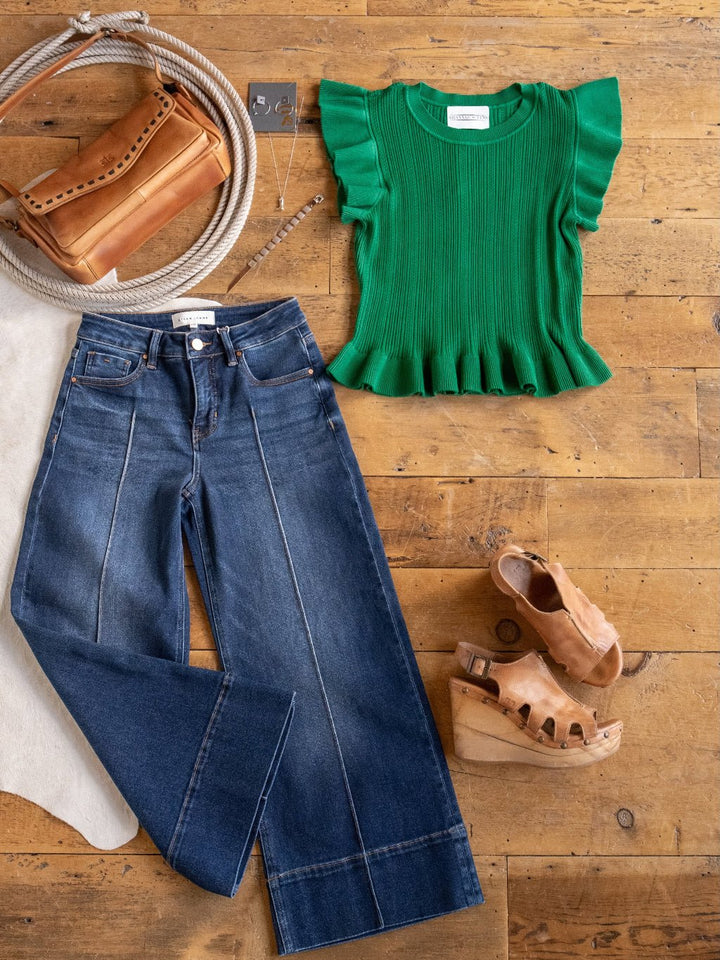 A flat lay displays the Sebring Emerald Flutter Sleeve Top by Savannah Sevens western life{&}style, paired with wide-leg blue jeans, tan platform sandals, a brown shoulder bag, coiled rope, and silver jewelry on a wooden floor.